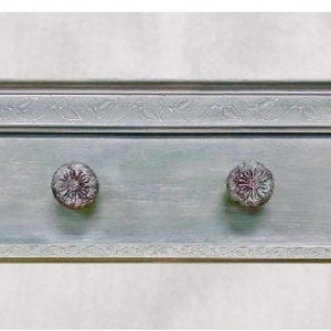 May include: A light blue wooden wall mounted coat rack with four decorative metal hooks. The hooks are round and have a floral design. The rack has a decorative trim around the top and bottom.
