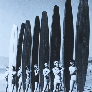 May include: A group of women in vintage swimwear stand behind a row of long surfboards on a beach. The women are smiling and holding onto the surfboards. The surfboards are all different colors and have a classic design.
