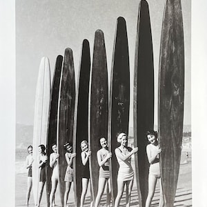 May include: A group of women stand on a beach holding long wooden surfboards. They are all wearing swimsuits and smiling. The surfboards are all different lengths and widths.