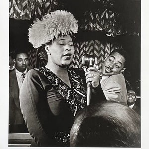 Puede incluir: Fotografía en blanco y negro de una mujer cantando en un micrófono. Lleva un gran sombrero de plumas y un vestido de lentejuelas. Un hombre está de pie detrás de ella, inclinado hacia el micrófono. El texto "WILLIAM P. GOTTLIEB The Golden Age of Jazz" está en la parte inferior de la imagen.