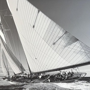 Könnte beinhalten: Schwarz-Weiß-Fotografie eines Segelbootes auf See, mit großen Segeln, die sich im Wind aufblähen. Das Boot ist mit Menschen besetzt, und das Wasser ist unruhig. Das Bild fängt die Bewegung und Kraft des Schiffes ein.