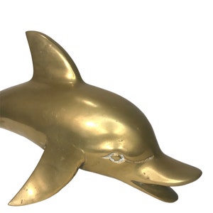 May include: A solid brass dolphin sculpture with a smooth, polished finish. The dolphin is depicted in a leaping pose with its mouth open.