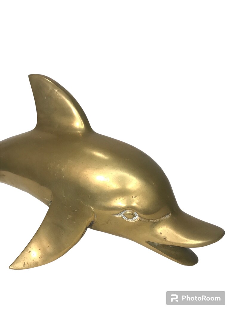 May include: A solid brass dolphin sculpture with a smooth, polished finish. The dolphin is depicted in a leaping pose with its mouth open.