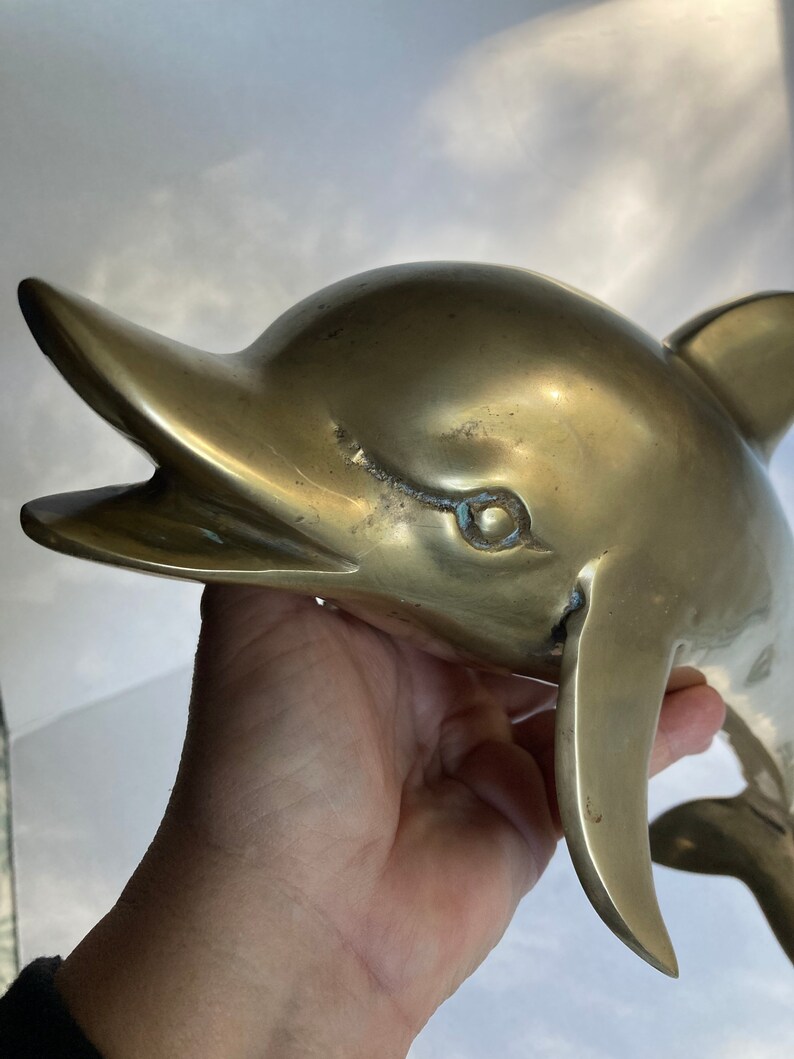 May include: A large, solid brass dolphin sculpture with a detailed, realistic design. The dolphin is depicted with its mouth open, as if it is about to leap out of the water.