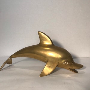 May include: A solid brass dolphin sculpture. The dolphin is in a leaping pose with its mouth open. The sculpture is a golden color.