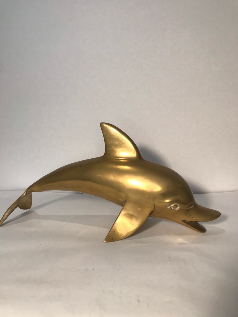 May include: A solid brass dolphin sculpture. The dolphin is in a leaping pose with its mouth open. The sculpture is a golden color.