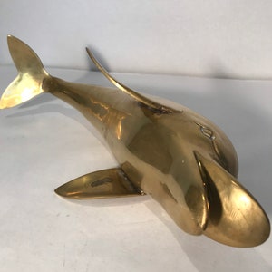May include: A solid brass dolphin sculpture with a smooth, polished finish. The dolphin is posed with its head tilted up and its tail fin extended.