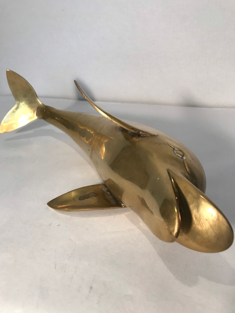 May include: A solid brass dolphin sculpture with a smooth, polished finish. The dolphin is posed with its head tilted up and its tail fin extended.