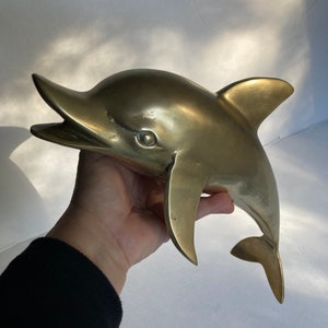 May include: A gold-colored brass dolphin sculpture. The dolphin is leaping out of the water with its mouth open and its tail fin extended.