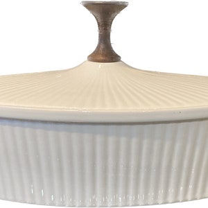 May include: Cream-colored oval casserole dish with a lid. The dish has vertical ribbed sides and a lid with a radial pattern. The lid is topped with a brown and gold handle. A kitchenware item.
