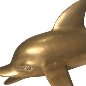 May include: A gold-colored brass dolphin sculpture. The dolphin is facing left and has a detailed, realistic design.