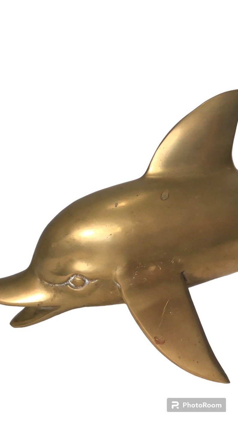May include: A gold-colored brass dolphin sculpture. The dolphin is facing left and has a detailed, realistic design.