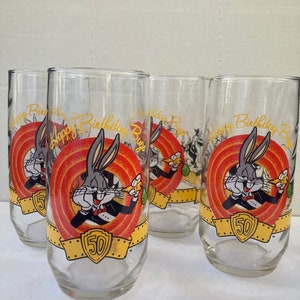 May include: Set of four clear glass tumblers with a Bugs Bunny 50th birthday design. Each glass features Bugs Bunny in a tuxedo holding a carrot, with the words "Happy Birthday Bugs" and "50" on a red and yellow banner.
