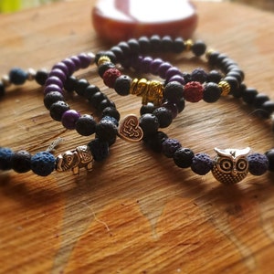 Puede incluir: Cuatro pulseras de cuentas con dijes decorativos. Las pulseras presentan cuentas de lava negras, cuentas moradas y dijes plateados y dorados, incluyendo un elefante, un búho y un dije en forma de corazón. Las pulseras se exhiben sobre una superficie de madera.