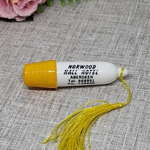 May include: A white thimble with a yellow textured top and a yellow tassel attached. The side of the thimble features black printed text that reads: NORWOOD HALL HOTEL ABERDEEN Tel: 868951 Tlx: 739440. The object is positioned near artificial flowers.