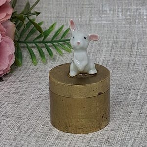 Caja de baratijas o pastillero de latón con un conejo de cerámica decorado con una flor rosa.