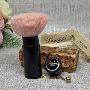 Op de afbeelding: Een vintage Lamkin Pixie poederdons met een zachte, lichtroze donskop bevestigd aan een zwarte cilindrische handgreep. De set bevat een kleine zwarte dop, een goudkleurige kralenketting en een rechthoekige doos met bloemmotieven en tekst.