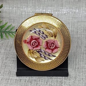 May include: A round gold-tone metal compact mirror featuring a textured border and a central decorative inlay. The inlay displays two pink roses with green leaves and small purple floral accents against a cream-colored background with a fine mesh pattern.
