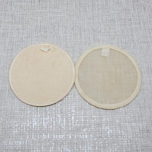 Puede incluir: Dos posavasos circulares de tela color blanco hueso con un centro liso y sólido y un borde reforzado ligeramente texturizado. Cada posavasos presenta una pequeña lengüeta de tela rectangular unida al borde superior del diseño.