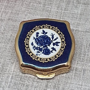 May include: A small, square, gold-toned metal compact case with rounded corners. The lid features a deep blue border surrounding a central white circular inlay decorated with a blue floral rose pattern and an intricate gold filigree frame.