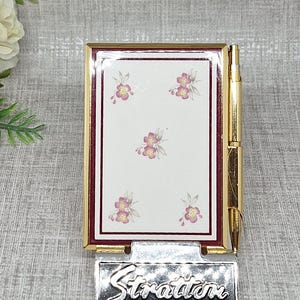 Puede incluir: Un estuche compacto rectangular de tono dorado de la marca Stratton que presenta un panel central blanco con un pequeño patrón floral rosa, un borde rojo oscuro y un bolígrafo dorado adjunto. La base tiene el nombre de la marca Stratton en plata.