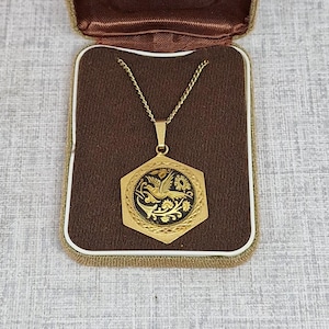 May include: A gold-coloured necklace with a hexagonal pendant featuring a black and gold design, displayed in an open, brown velvet-lined jewellery box. The pendant showcases a bird and floral motif. The box is beige with a brown interior.