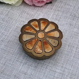 May include: A small, round, decorative box with a flower-like design. The box has a bronze-colored metal frame and a segmented, light brown and beige interior. The box is sitting on a gray fabric surface, with pink and yellow flowers in the background.