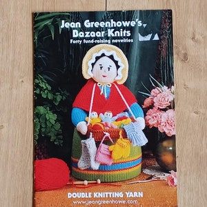 May include: A book cover featuring a colorful illustration of a knitted doll holding a basket of knitted items. The book title is "Jean Greenhowe's Bazaar Knits" and the subtitle is "Forty fund-raising novelties". The book cover also includes the text "Double Knitting Yarn" and a website address.