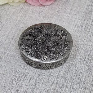 May include: A small, round, silver-toned trinket box with a detailed floral design on the lid. The flowers are intricately sculpted, and the box has a textured, patterned band around the sides. The box is sitting on a light gray surface.