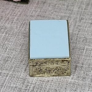 Peut inclure: Une petite boîte rectangulaire dotée d'un couvercle supérieur bleu clair lisse et d'une base métallique avec un motif floral complexe en relief sur les côtés. La boîte est placée près d'un bouquet de fleurs artificielles roses et crème.