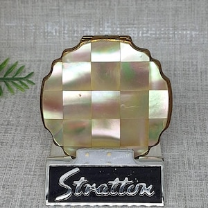 May include: A round compact mirror featuring a checkered mother-of-pearl inlay on the lid. The base is silver-toned metal with a black rectangular plaque displaying the word Stratton in silver script lettering. The item has a scalloped edge.
