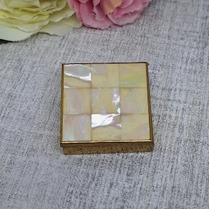 May include: A small, square, vintage compact with a gold-colored metal frame. The top is inlaid with iridescent mother-of-pearl tiles. The compact is closed and sits on a textured surface, with pink and yellow flowers in the background.