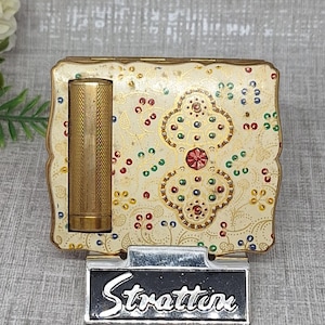 May include: A square Stratton compact case with a cream-colored enamel finish featuring a delicate floral pattern in red, green, and blue dots. A gold-toned cylindrical lipstick holder is attached to the left side, and a black plaque with the white text Stratton is positioned at the base.