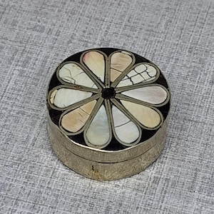 May include: A small, round, metallic trinket box featuring a lid decorated with a floral inlay pattern. The design consists of ten petal-shaped pieces of iridescent, cream-colored shell set against a dark background, forming a flower shape.