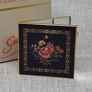 Puede incluir: Un espejo compacto cuadrado con marco de metal dorado y tapa azul oscuro con un estampado de ramo de flores. El espejo se muestra junto a su caja de cartón beige original, que lleva impreso el texto EXCLUSIVE ACCESSORIES.