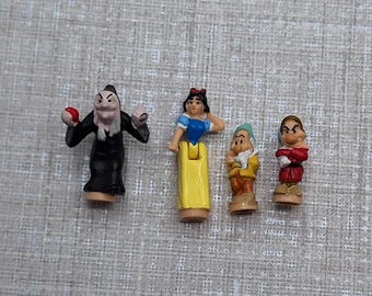 Può includere: Figurine in miniatura dei personaggi di Biancaneve. Le figure includono la Regina Cattiva, Biancaneve e due dei sette nani. La Regina Cattiva indossa una veste nera, Biancaneve un vestito giallo e i nani vari abiti.