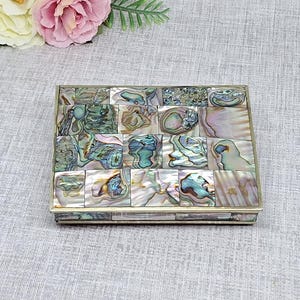 Peut inclure: Une boîte rectangulaire dotée d'un couvercle en mosaïque composé de carreaux de nacre d'ormeau irisés dans des tons de vert, bleu et rose. La boîte présente un cadre métallique et une surface plane, mettant en valeur la texture naturelle nacrée.