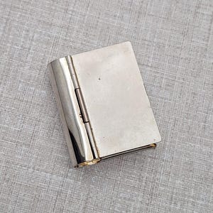 May include: A small, silver-coloured metal box with a hinged design, resembling a miniature book. The box is rectangular with rounded corners and a shiny, reflective surface. The hinge is visible on the left side, and the edges have a gold-coloured trim.