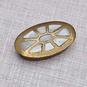 Könnte beinhalten: Ein ovaler Knopf mit einem metallischen, messingfarbenen Rahmen in verwitterter Optik. Die Mitte des Knopfes ist mit weißen, schillernden Perlmuttsegmenten eingelegt, die in einem strahlenförmigen, radartigen Muster um ein zentrales Oval angeordnet sind.