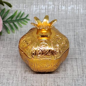 Pode incluir: Uma caixa de metal dourado em forma de romã com um padrão intrincado de volutas em relevo em toda a sua superfície e uma tampa com um remate em forma de coroa estilizada, posicionada ao lado de um ramo de rosas artificiais rosa e creme.
