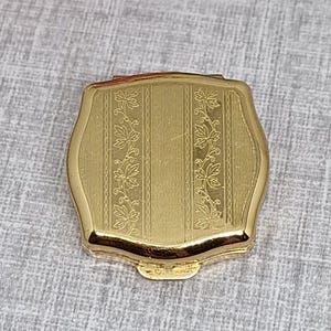 May include: A gold-toned metal compact case with a square shape and rounded corners. The surface features a central vertical panel flanked by two decorative bands of engraved leaf patterns, all set against a fine, textured, brushed metal finish.