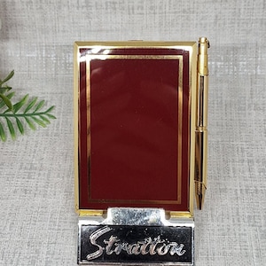 Puede incluir: Un soporte rectangular para bloc de notas en color burdeos con un marco de metal dorado y un bolígrafo a juego en tono dorado sujeto al lateral. La base presenta una placa de metal negro con la palabra Stratton grabada en una fuente cursiva plateada.