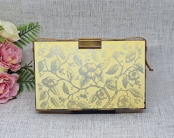 May include: A vintage gold-toned clutch with a floral pattern. The rectangular purse has a clasp closure and a small handle. A bouquet of pink and yellow flowers sits to the left of the purse.