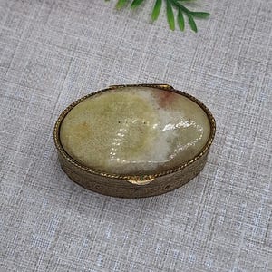 May include: An oval antique trinket box with a pale green and white stone top. The box has a gold-coloured metal frame with detailed patterns around the edges. The box is closed and rests on a neutral-coloured surface.