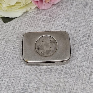 Puede incluir: Una caja rectangular de metal plateado con un diseño circular texturizado en el centro. La caja tiene una apariencia ligeramente desgastada, lo que sugiere edad o uso. El fondo presenta una superficie gris texturizada.