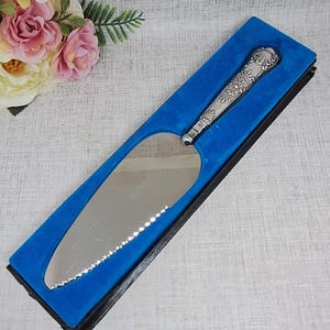 Puede incluir: Un servidor de pastel plateado con un mango decorativo, presentado en una caja forrada de terciopelo azul. El servidor tiene un borde dentado y detalles florales en el mango. Un ramo de flores rosas y crema está en el fondo.