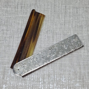 Könnte beinhalten: Ein klappbarer Taschenkam mit einem braunen, schildpattfarbenen Kunststoffkamm und einem silberfarbenen Metallgriff mit geprägtem Blumenmuster. Der Kamm ist teilweise ausgeklappt und zeigt die Zinken sowie das dekorative Metallgehäuse.