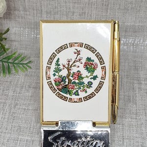 May include: A rectangular Stratton compact mirror with a white enamel cover featuring a circular floral design in pink, green, and brown. The compact has a gold-tone metal frame, a side-mounted gold pen, and a black base with the word Stratton in silver.