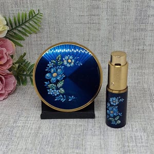 May include: A vintage blue compact and perfume bottle set. The compact is round with a gold rim and a floral design. The perfume bottle is cylindrical with a gold top and matching floral details. The set is displayed on a neutral surface.