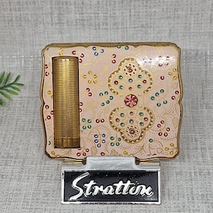 May include: A square powder compact with a pale pink enamel finish, featuring gold-tone filigree patterns and colorful embedded rhinestones. A textured gold-tone lipstick tube is attached to the left side. The compact rests on a black stand with the white text Stratton.